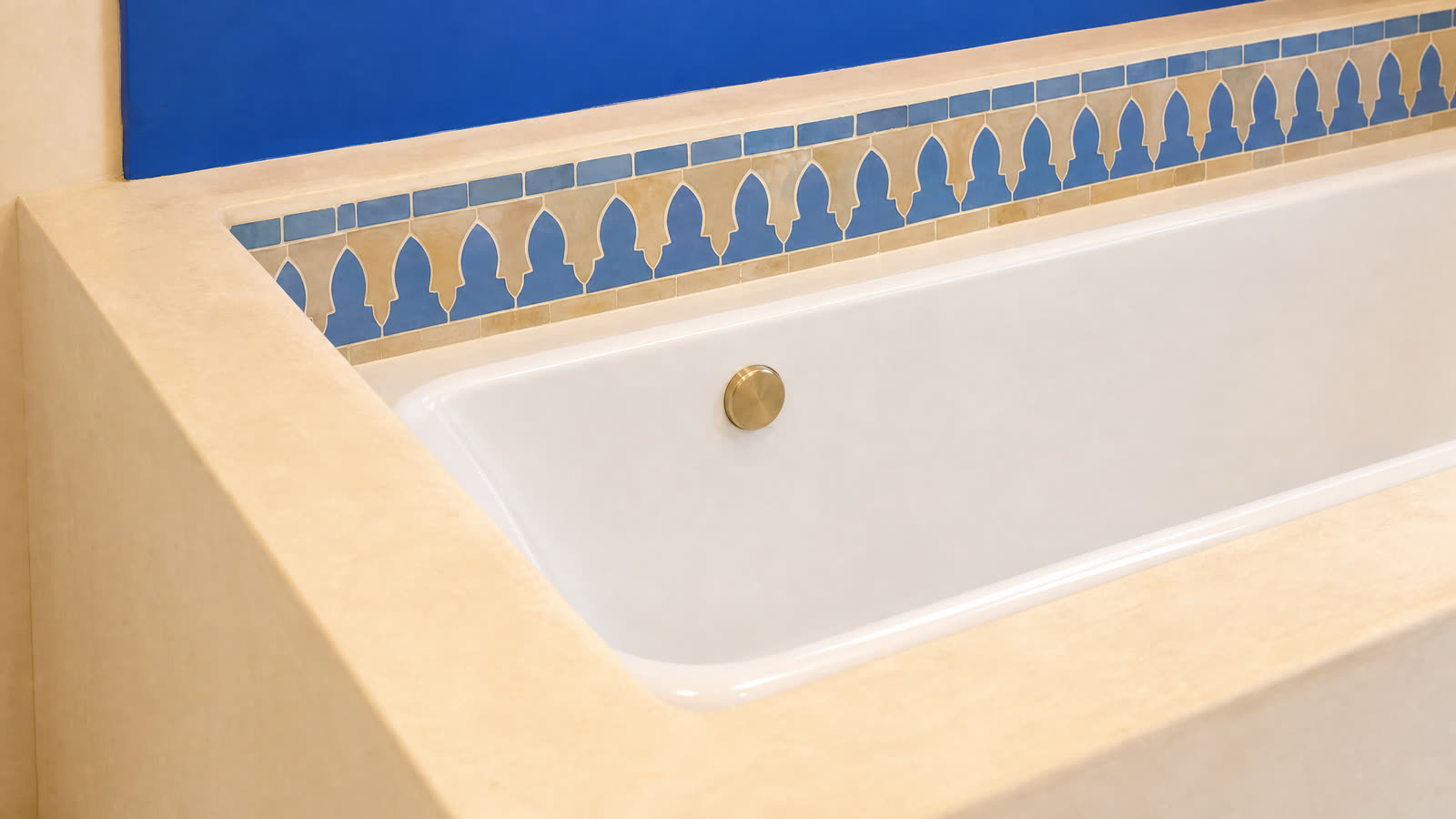 A blue and gold zellige frieze around the tub, on a sand-coloured plaster.