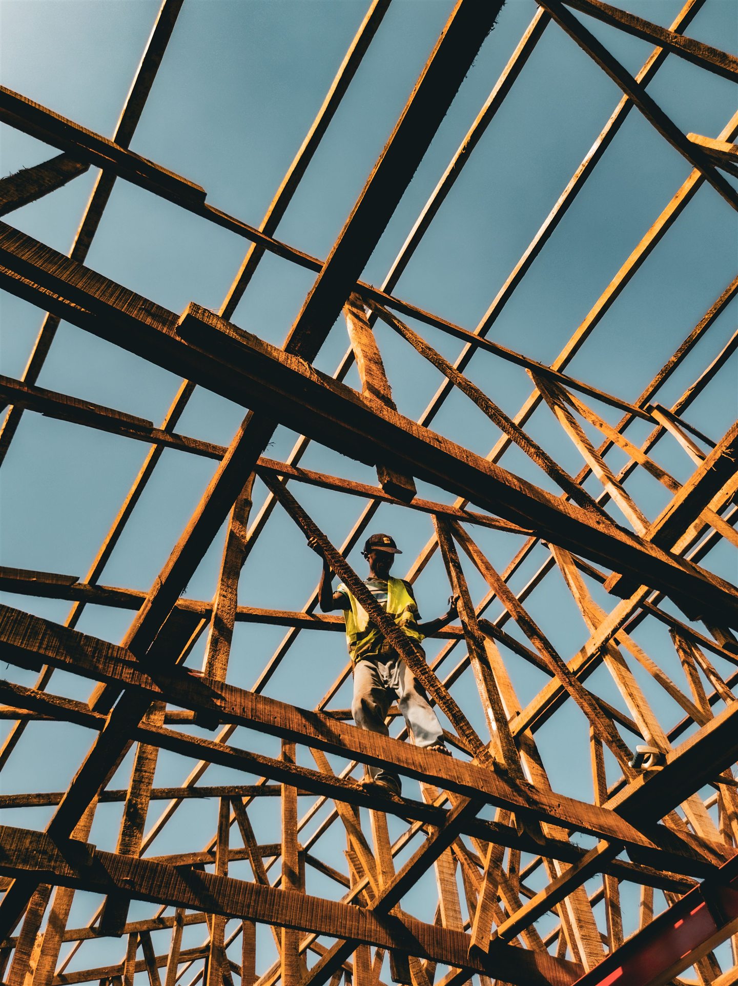 Carpenter in a yellow hard hat among the roof trusses, against a blue sky