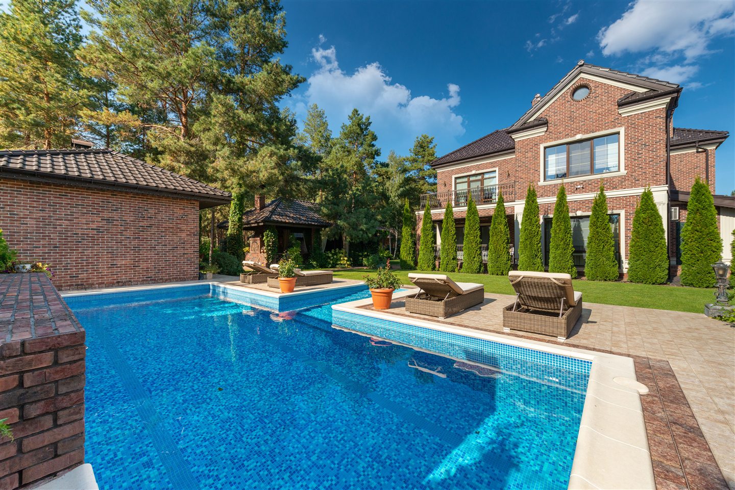 Piscine en mosaïque bleue bordée de chaises longues, devant une maison de brique entourée de cèdres et de pins