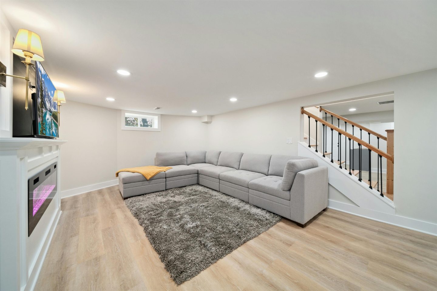 Salle familiale au sous-sol avec grand sofa sectionnel, foyer électrique, plancher clair et escalier à barreaux