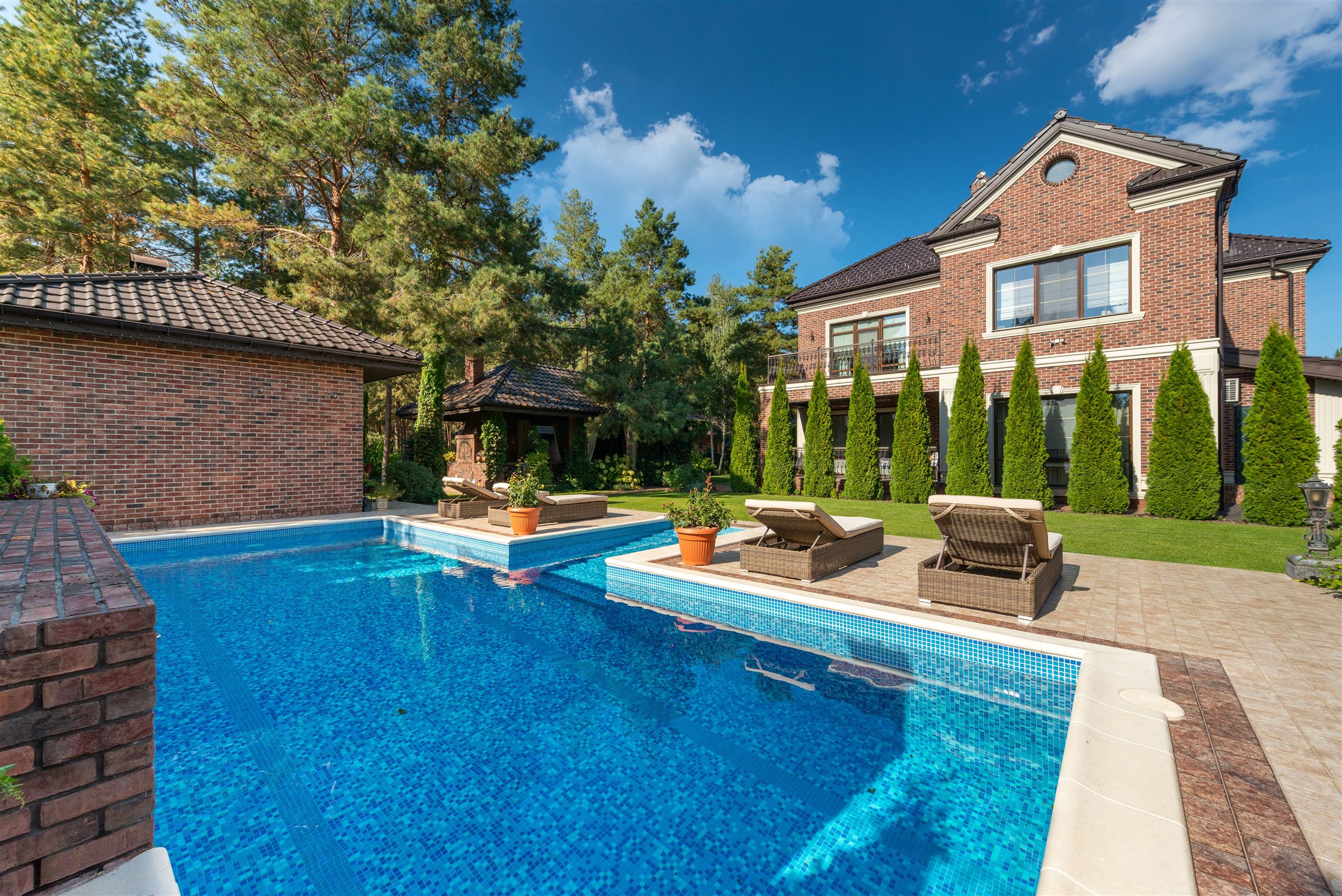 Piscine en mosaïque bleue bordée de chaises longues, devant une maison de brique entourée de cèdres et de pins