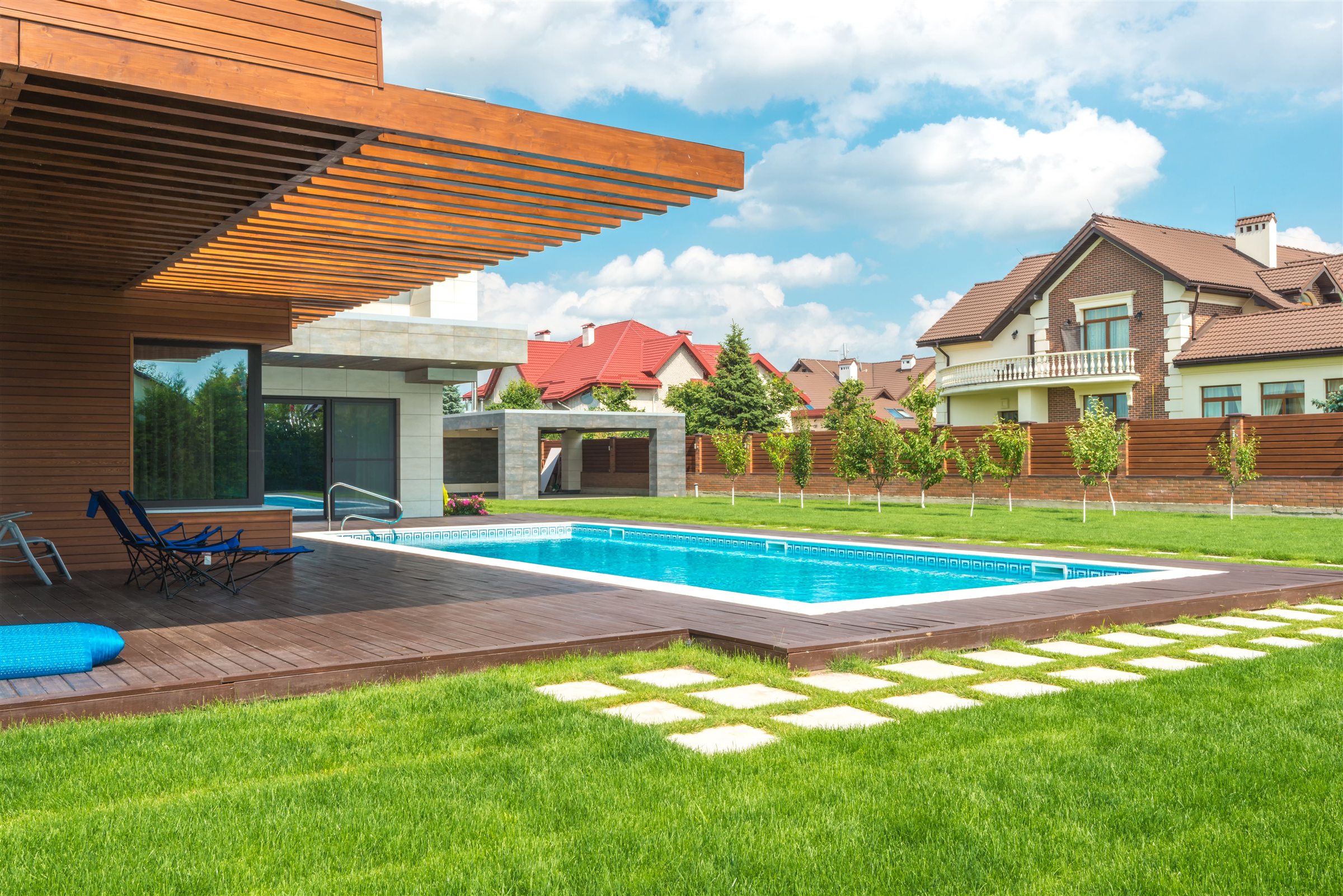 In-ground pool with a wooden deck under a pergola, lawn and stepping stones